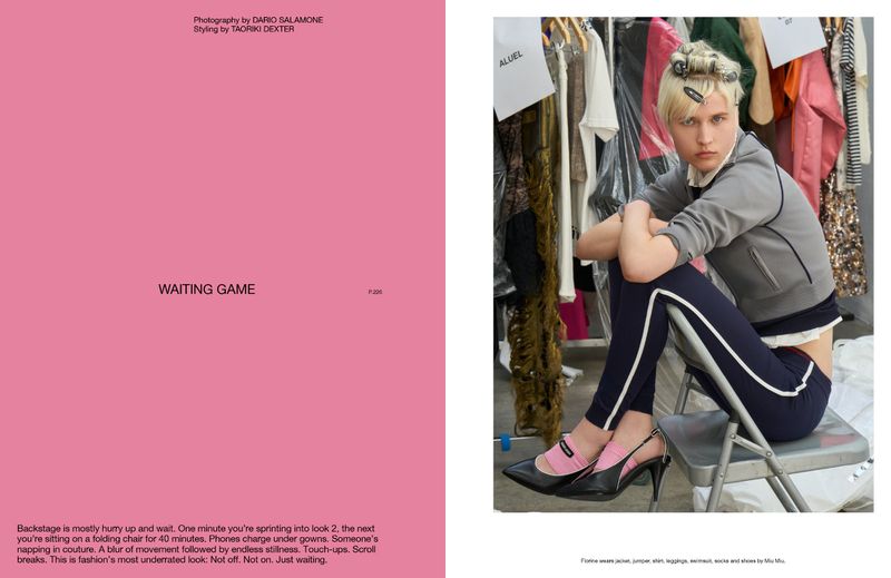 Blonde fashion model in gray jacket and leggings poses on folding chair in a photo studio with wardrobe rack in the background.