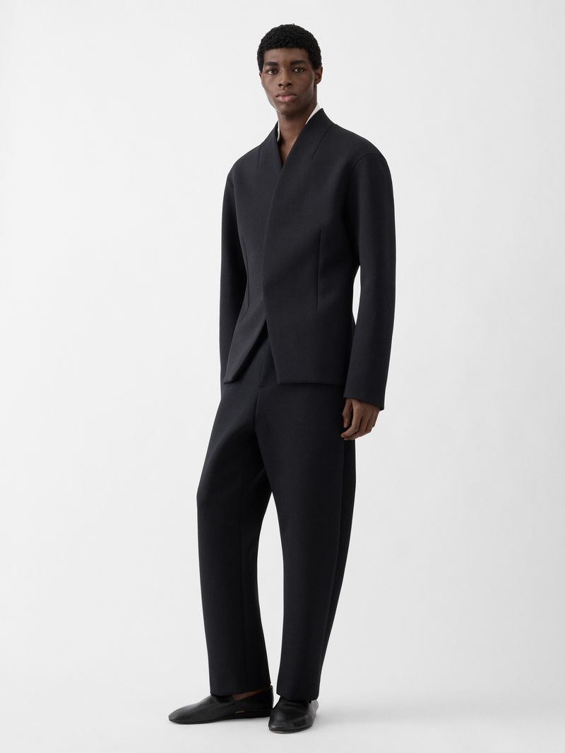 Model Zaram Obasi wearing a sculptural black suit and black loafers against a seamless white studio background for Jacquemus S/S 26.