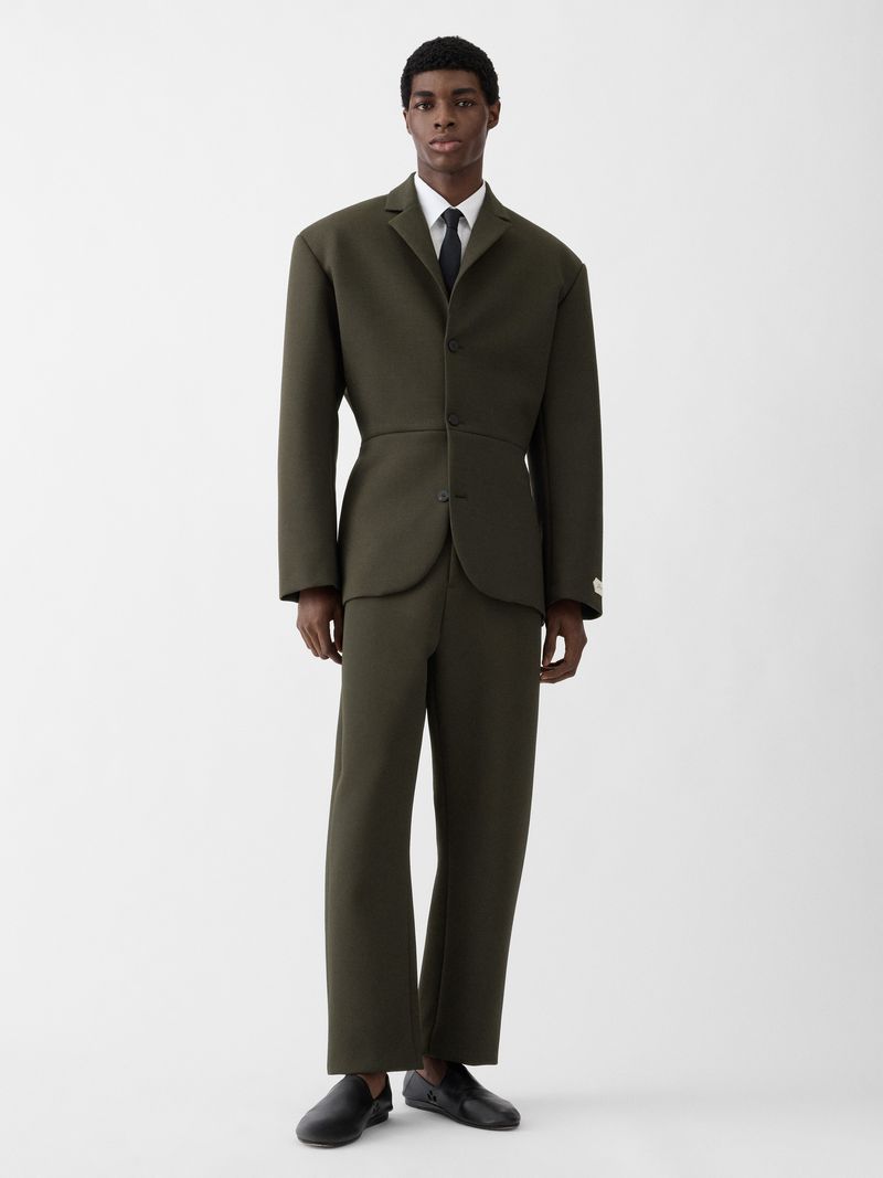 Model Zaram Obasi wears a structured olive green tailored suit with a white shirt and black tie against a white background.