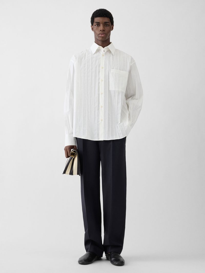 Model Zaram Obasi standing in a white pleated button-down shirt and black tailored trousers against a grey studio backdrop.