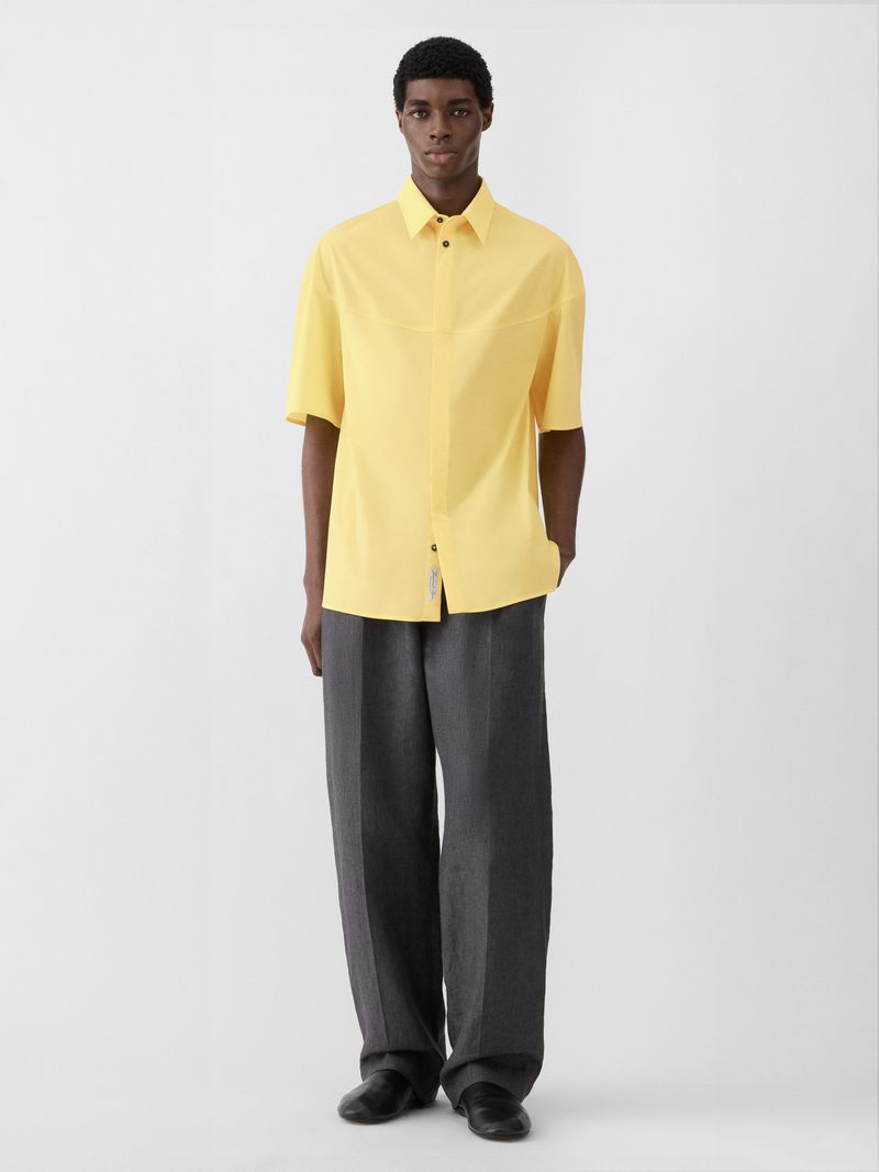 Model Zaram Obasi wearing a butter yellow short-sleeve shirt and charcoal wide-leg trousers against a light grey studio backdrop.