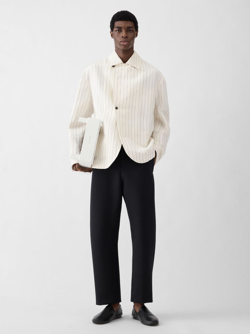 Model Zaram Obasi wearing a cream striped jacket and black trousers holding a white handbag against a white background.