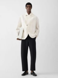 Model Zaram Obasi wearing a cream striped jacket and black trousers holding a white handbag against a white background.