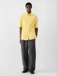 Model Zaram Obasi wearing a butter yellow short-sleeve shirt and charcoal wide-leg trousers against a light grey studio backdrop.