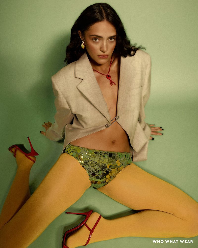 Fashion editorial shot of a woman in a beige blazer, sequin shorts, and yellow tights sitting against a green background in studio light.
