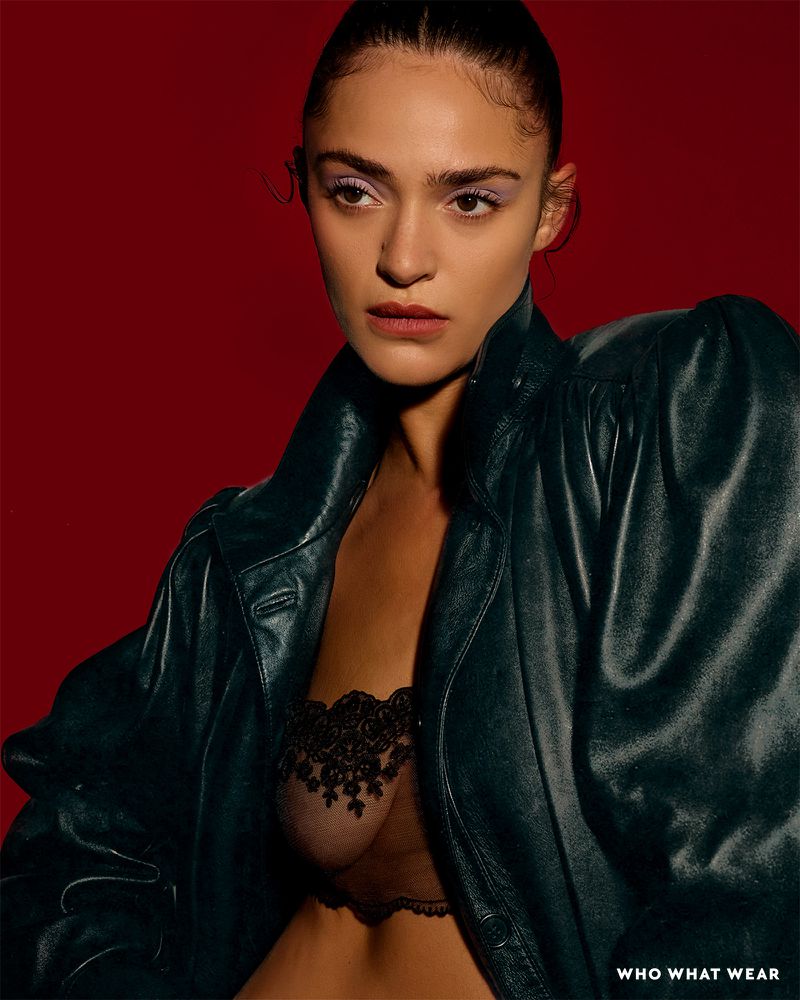 Portrait of a woman with light skin wearing a black leather jacket and lace bra against a red background in a fashion-editorial style photograph.