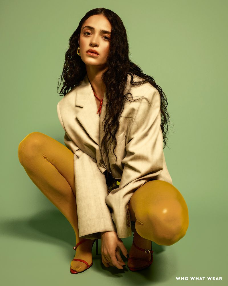 A woman with long dark hair is crouched down, wearing a tan blazer, a red necklace, yellow tights, and strappy heels against a green background.