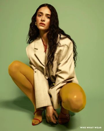 A woman with long dark hair is crouched down, wearing a tan blazer, a red necklace, yellow tights, and strappy heels against a green background.