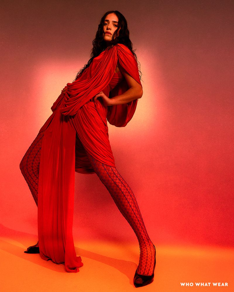 A long-haired female fashion model strikes a pose in a dramatic, draped red dress and patterned tights against a blended gradient background.
