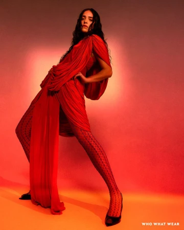 A long-haired female fashion model strikes a pose in a dramatic, draped red dress and patterned tights against a blended gradient background.