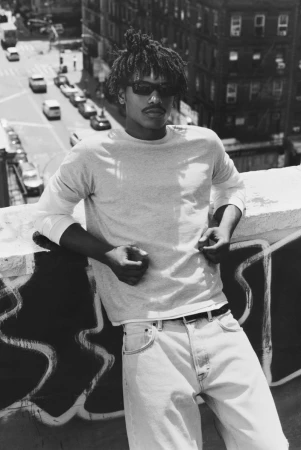 Black man in dark sunglasses, long-sleeve tee, and light jeans leaning against a rooftop ledge with city streets below.