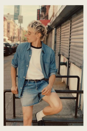 Model in denim shirt and denim shorts leaning against a black metal railing on a city street.