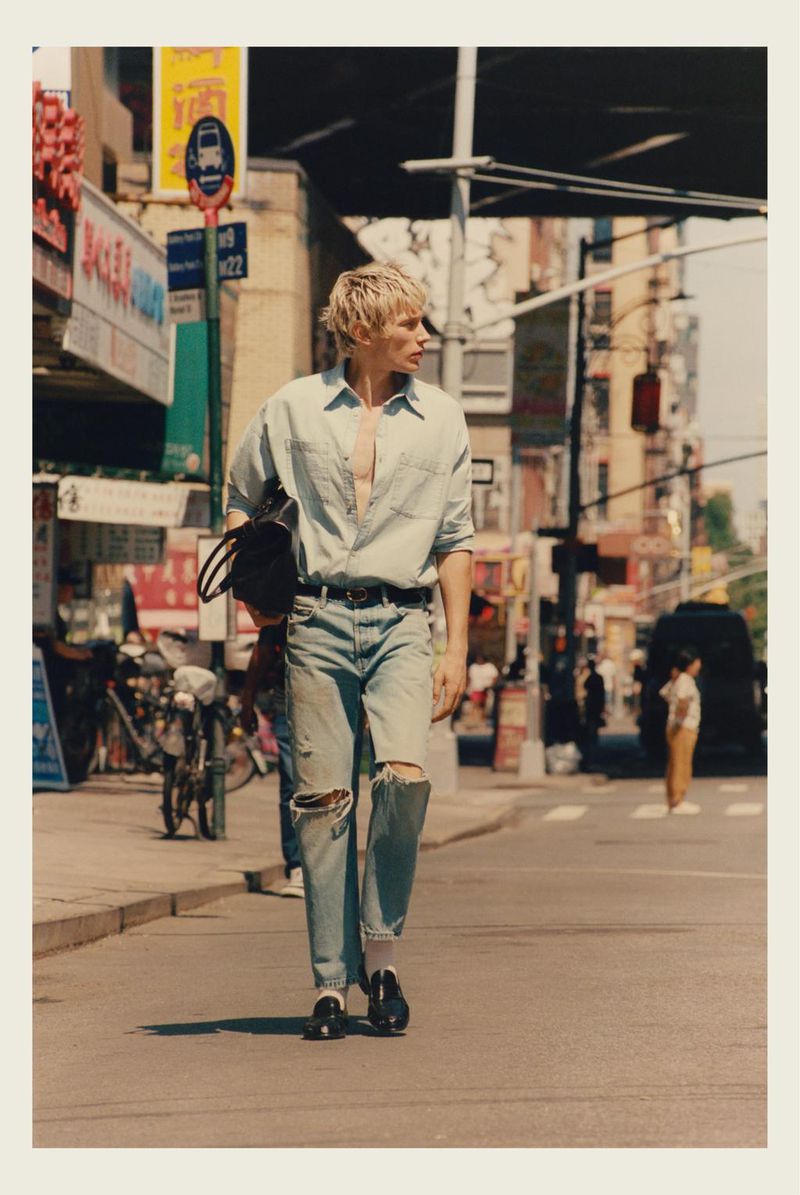 Model wearing a light-wash denim shirt and distressed jeans walking down a sunny urban street with city traffic in the background.