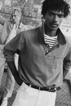 Black and white portrait of a black man wearing a denim shirt and white trousers standing outdoors against a wire fence
