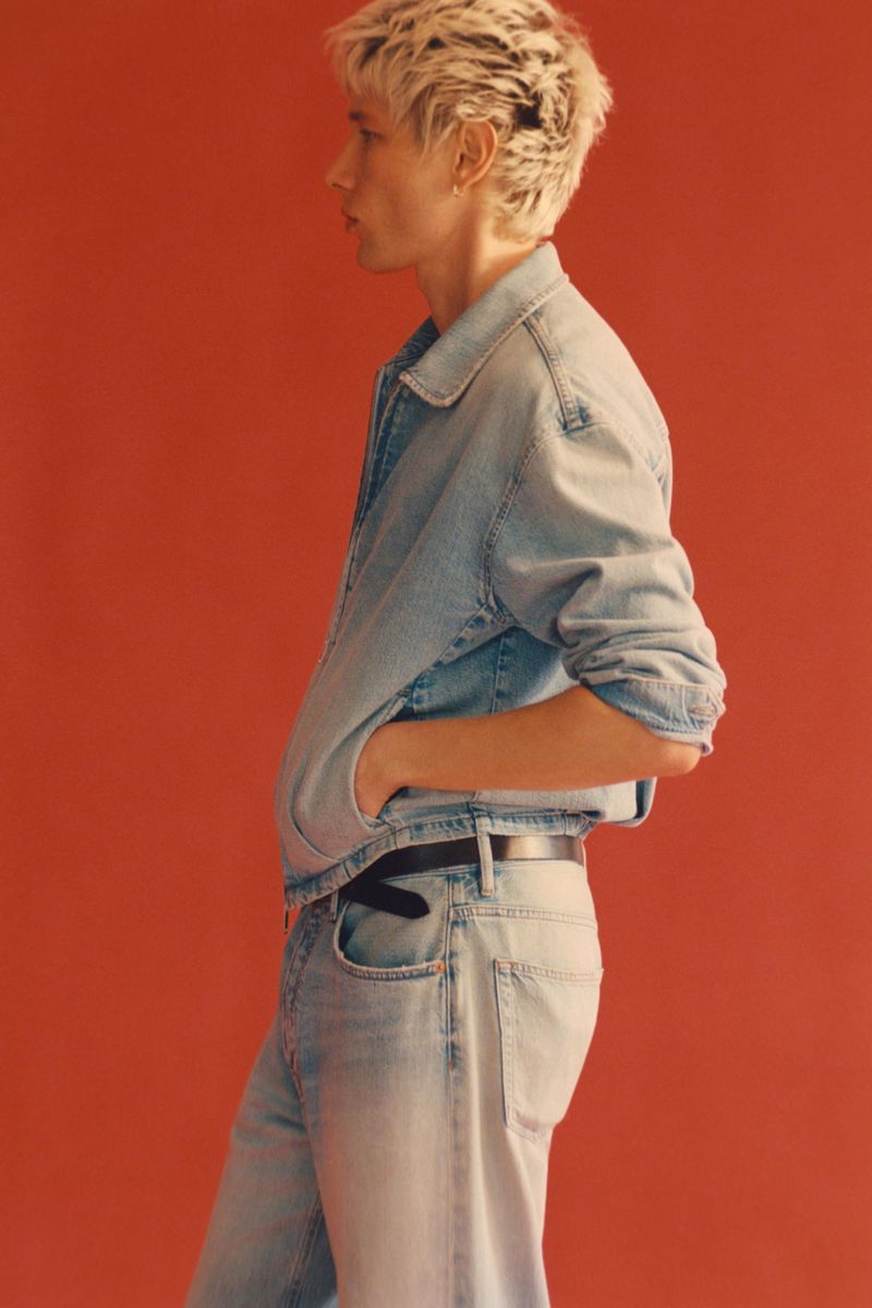 Model with short bleached blonde hair in profile wearing a light-wash denim jacket and matching jeans against a solid red backdrop.