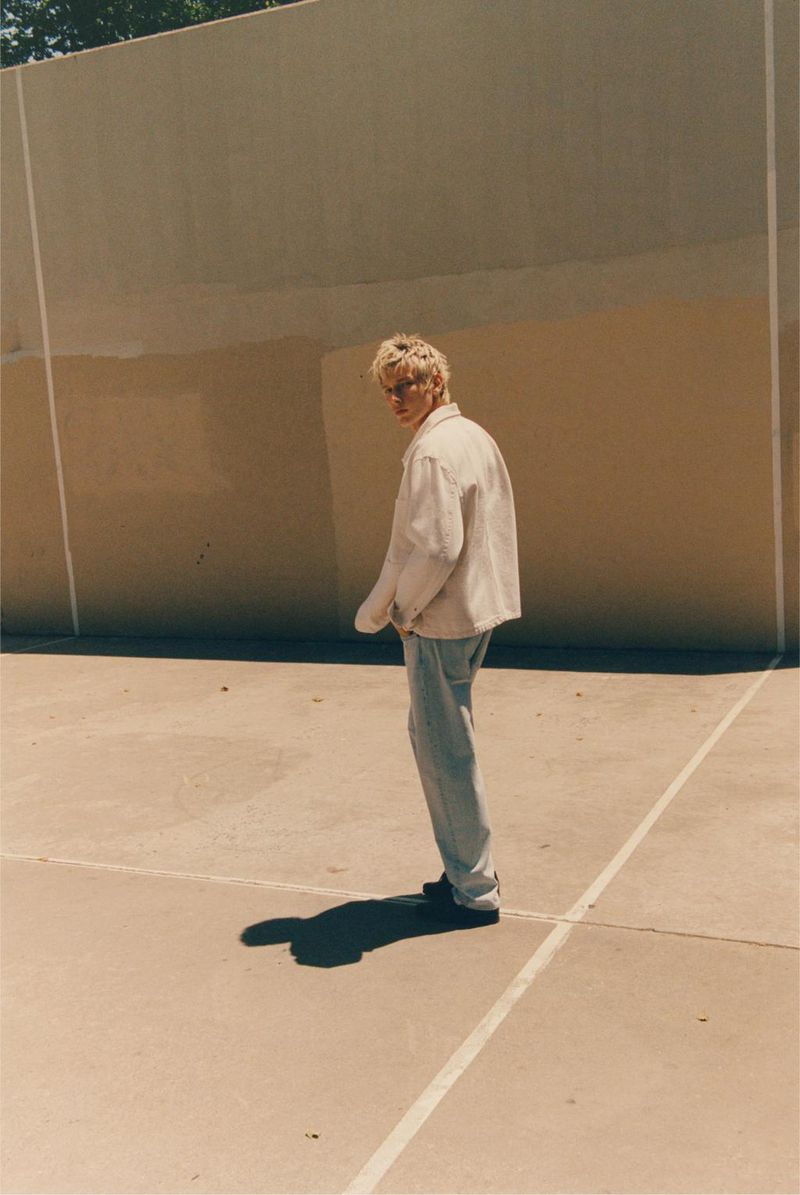 Man in a light cream jacket and pale denim jeans standing on an outdoor concrete court in direct sunlight.