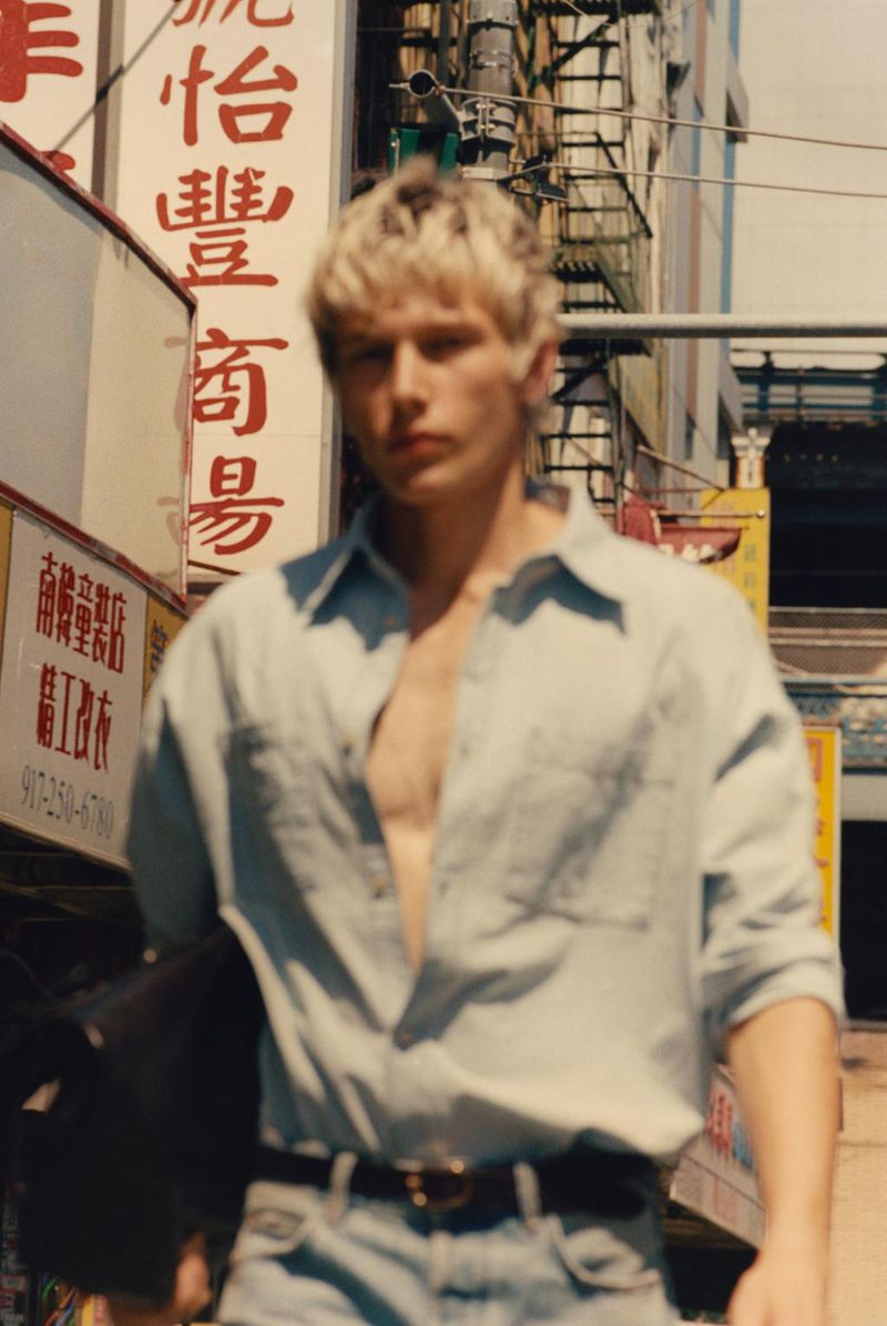 Model walking down a sunlit city street wearing an unbuttoned light blue denim shirt and matching jeans.
