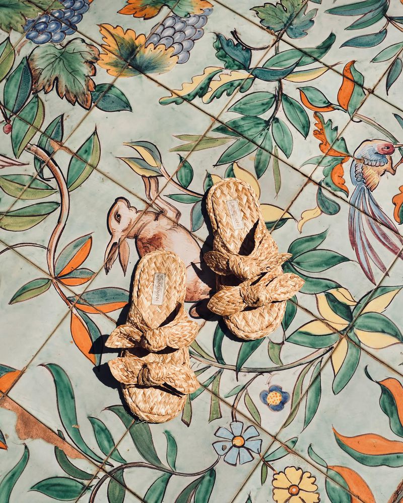 Pair of woven raffia bow slide sandals resting on ornate painted ceramic floor tiles under bright sunlight.