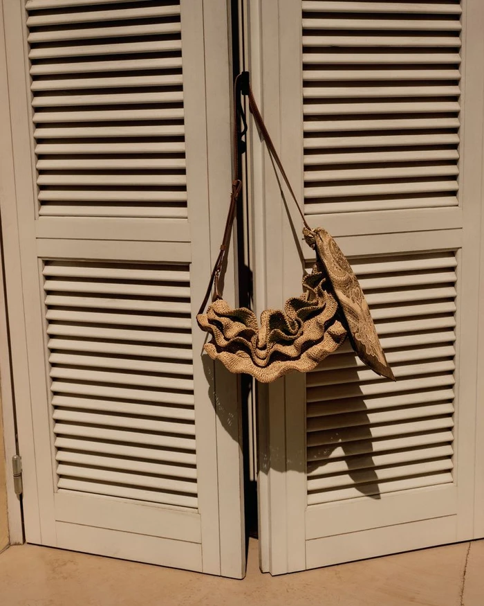 Woven straw ruffled shoulder bag hanging from white louvered wooden shutters in bright direct sunlight.