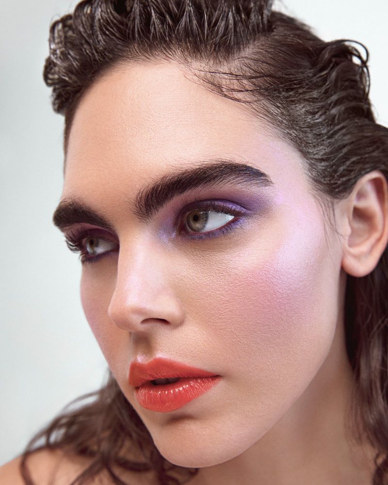 Close-up beauty portrait of a woman with bold brows, purple eyeshadow, and coral lipstick against a plain light background.