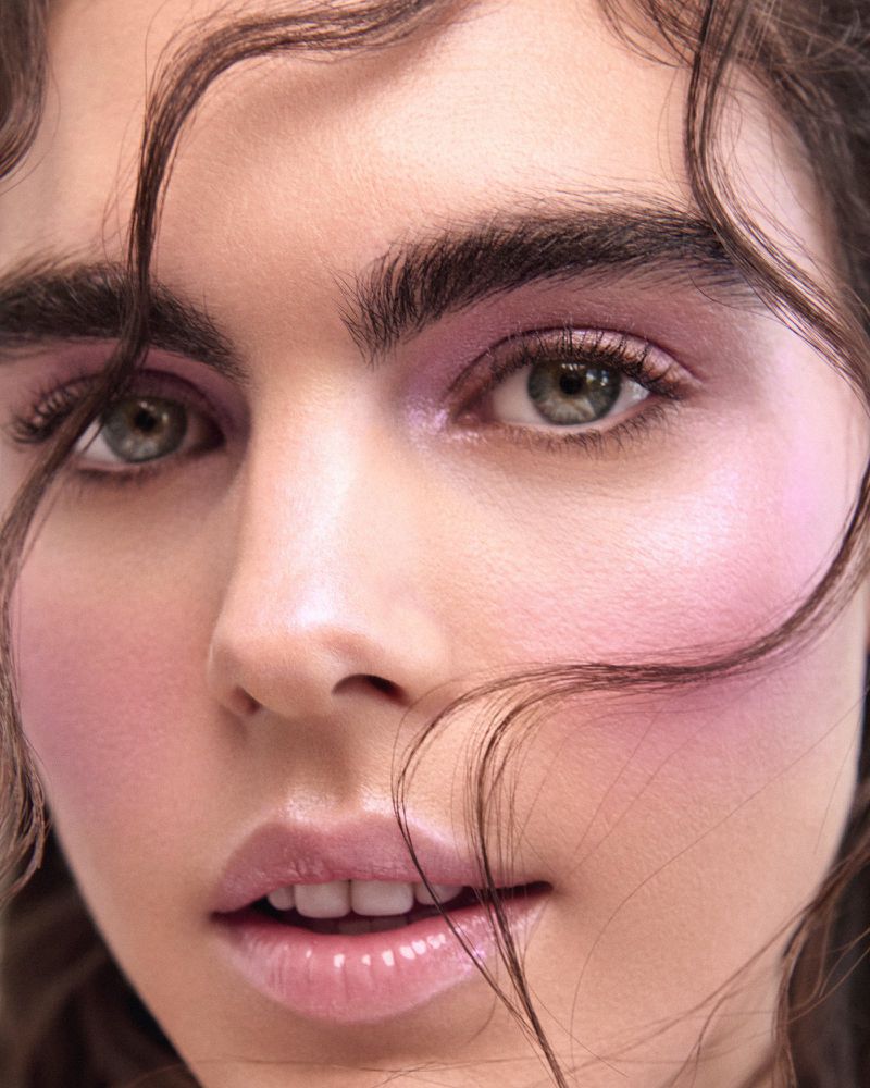 Close-up beauty portrait of a woman with lilac eyeshadow, pink blush, bold eyebrows, and wisps of hair across her face.