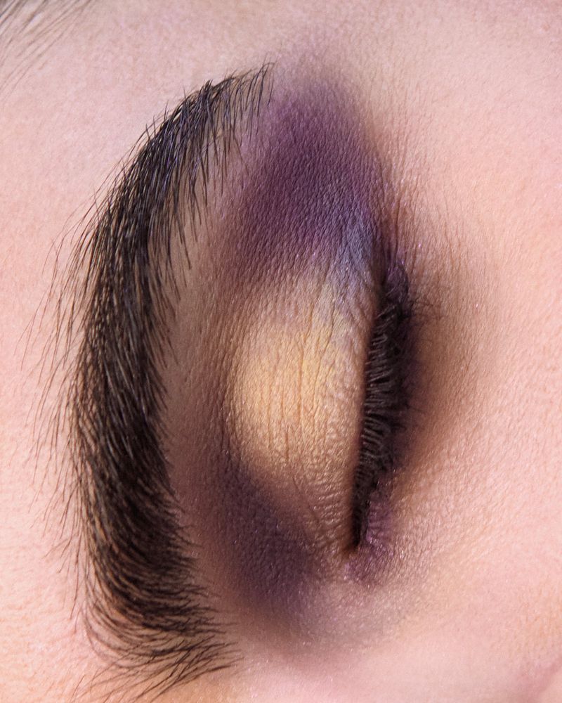 Extreme close-up of a closed eye styled with blended purple and yellow eyeshadow makeup and a dark defined eyebrow.