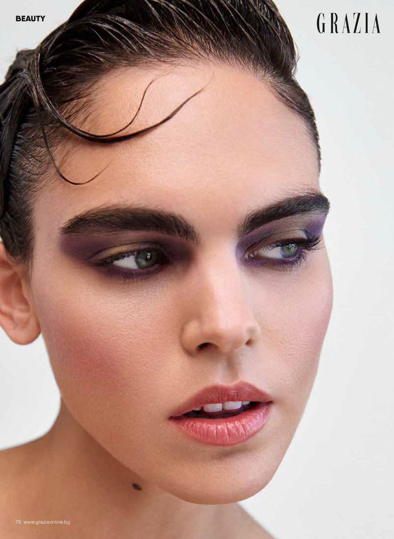 Close-up beauty portrait of a woman wearing dramatic purple eyeshadow with wet-look hair against a clean white backdrop.