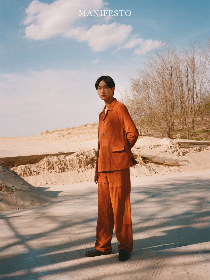 Model Hyun Jun Kim stands in a rust-orange suit on a bright sandy outdoor landscape under a blue sky for Manifesto Magazine.