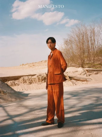 Model Hyun Jun Kim stands in a rust-orange suit on a bright sandy outdoor landscape under a blue sky for Manifesto Magazine.
