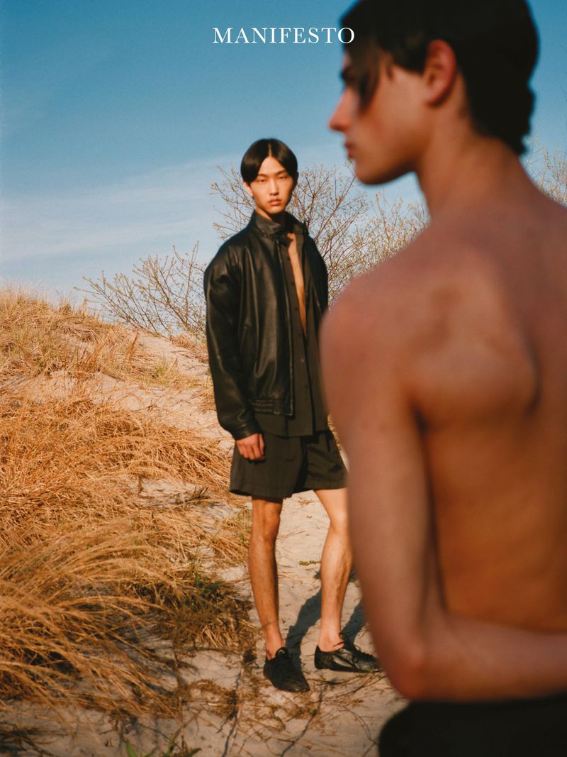 Two male models on a sandy dune under a clear sky, one shirtless in the foreground and one wearing a black leather jacket