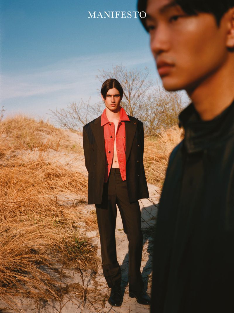 Fashion editorial photo of a male model wearing an open red shirt and black suit standing on grassy dunes under daylight.
