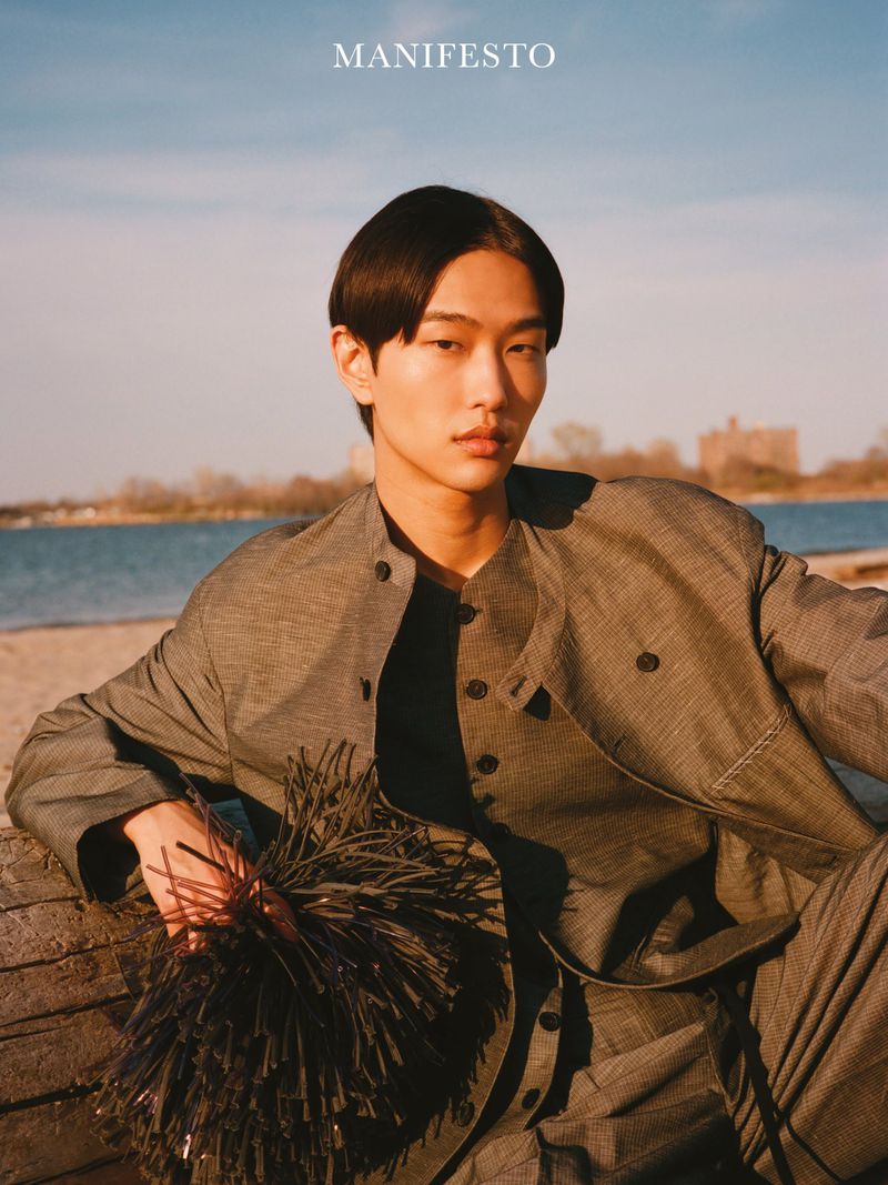 Asian man in a grey buttoned jacket seated outdoors by a body of water holding a dark textured prop for Manifesto Magazine