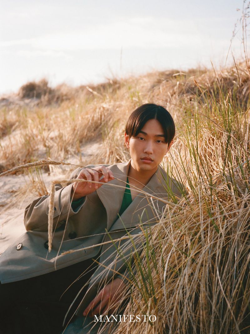 Model Hyun Jun Kim seated among dry tall grass on sand dunes wearing a beige trench coat for Manifesto Magazine.