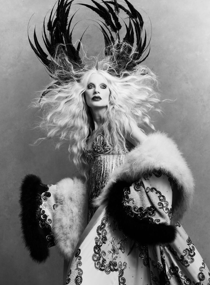 Model Kristen McMenamy wearing an elaborate black feather headdress and fur-trimmed embroidered gown in a black and white portrait.