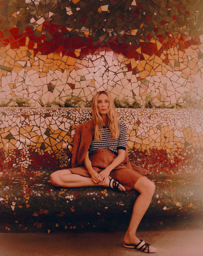 A blonde woman sits cross-legged on a mosaic bench in a striped shirt, brown shorts, and sandals, with a brown jacket on her shoulder.