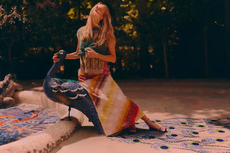Blonde woman in knitwear seated on a mosaic peacock sculpture in an outdoor garden with dappled light and trees in the background.