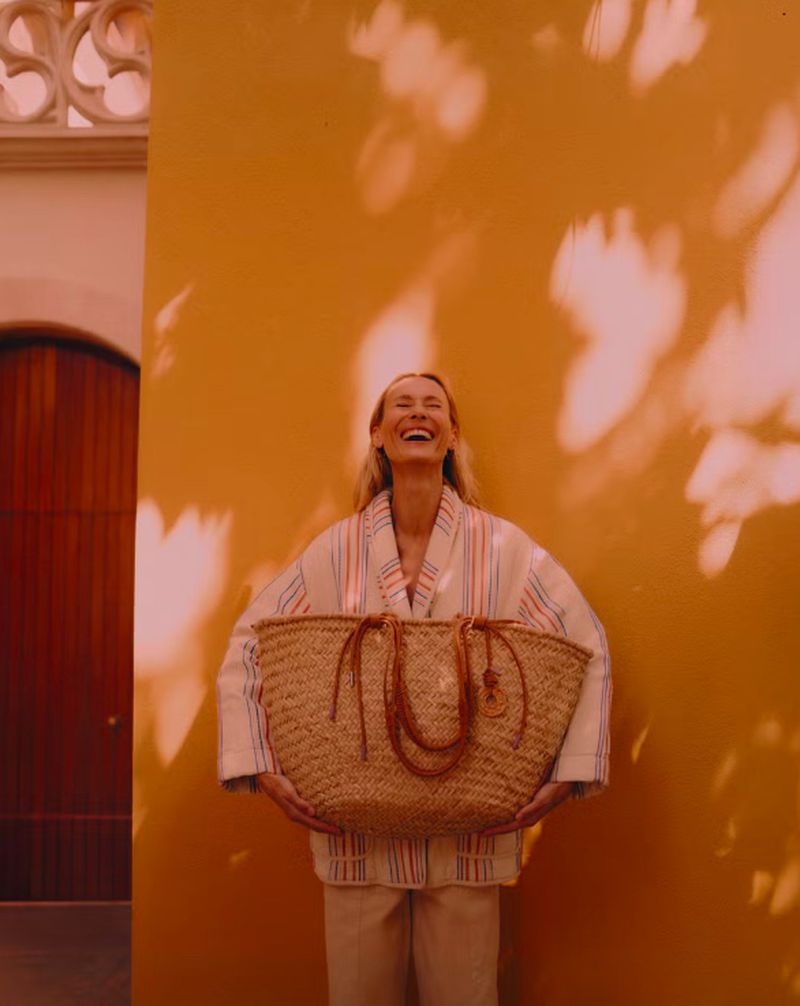 Smiling woman in a striped robe holding a woven market basket against an orange wall with shadow patterns, creating a warm, joyful scene.