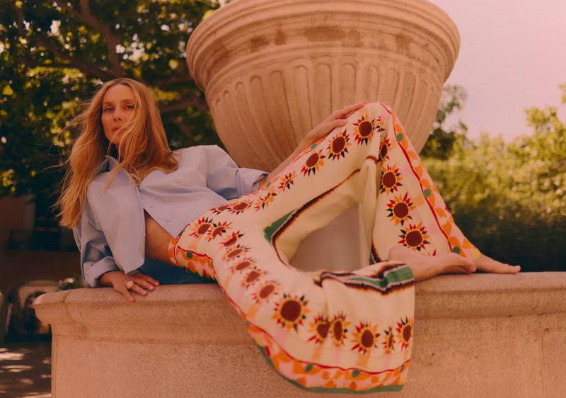 A woman in a light blue shirt and patterned pants reclines against a stone structure in an outdoor setting with natural light.