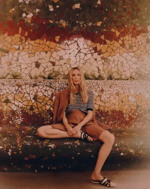 Vanesa Lorenzo seated cross-legged on a mosaic tile bench wearing a brown blazer, striped top, brown shorts, and slide sandals.