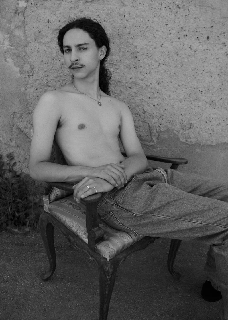 Shirtless man with dark hair and mustache seated in an antique wooden armchair against a textured concrete wall.