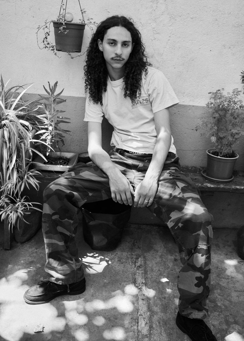 A young man with long curly hair seated outdoors wearing a white t-shirt and camouflage cargo pants surrounded by potted plants.
