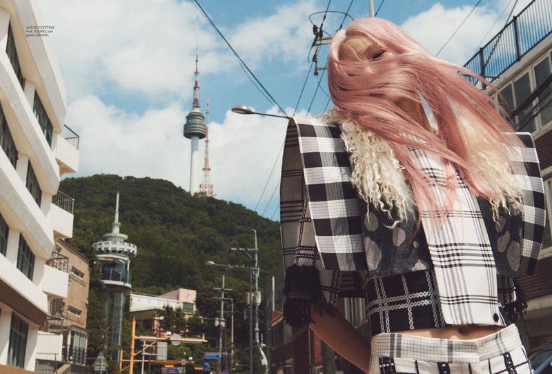Model Hoyeon Jung with windblown pink hair wearing a black and white plaid structured top with N Seoul Tower in the background.