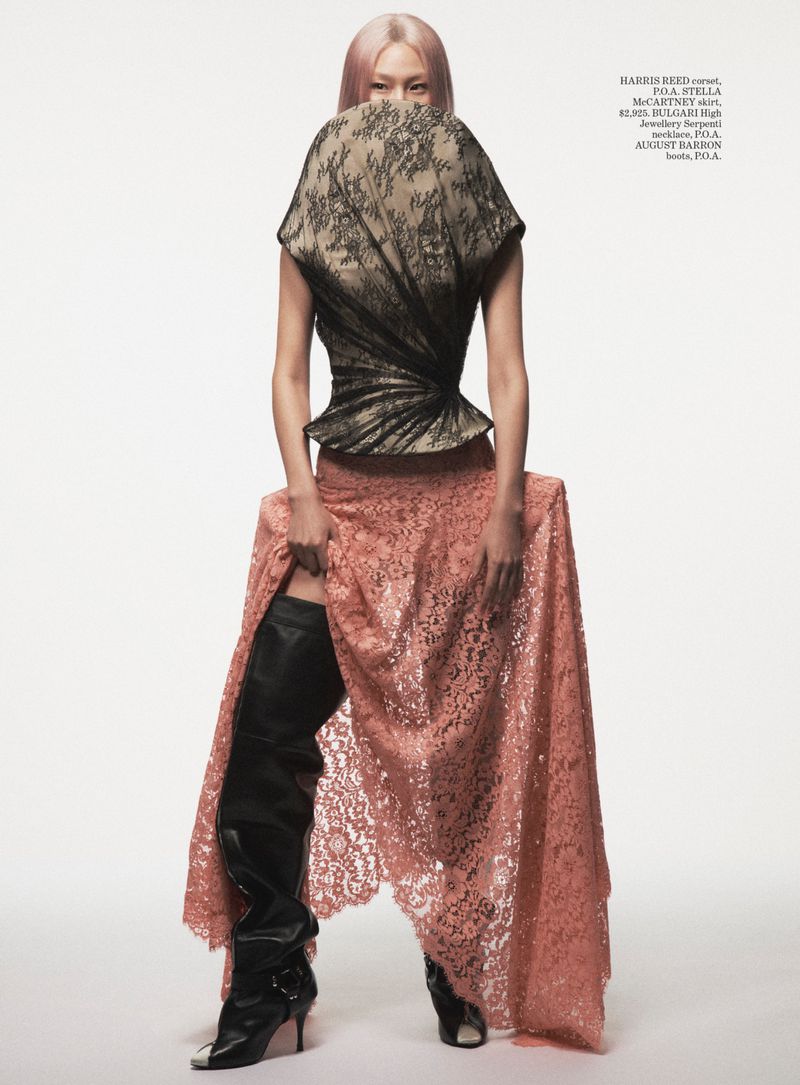 Hoyeon Jung posing in a high sculptural lace top, peach lace skirt, and black thigh-high boots against a white studio backdrop.