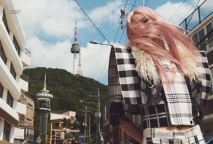 Model Hoyeon Jung with windblown pink hair wearing a black and white plaid structured top with N Seoul Tower in the background.