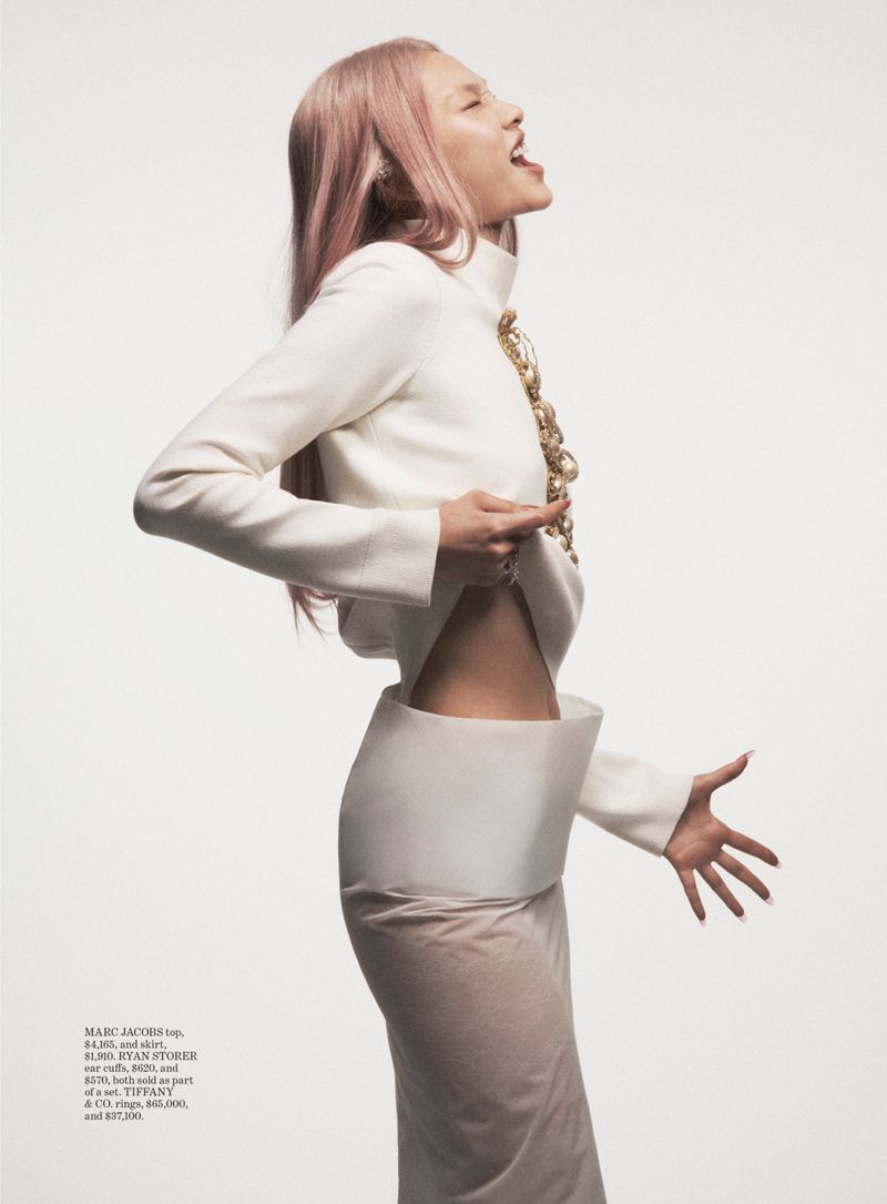 Model Hoyeon Jung with pink hair poses in profile wearing a sculptural white top and skirt against a bright white background.