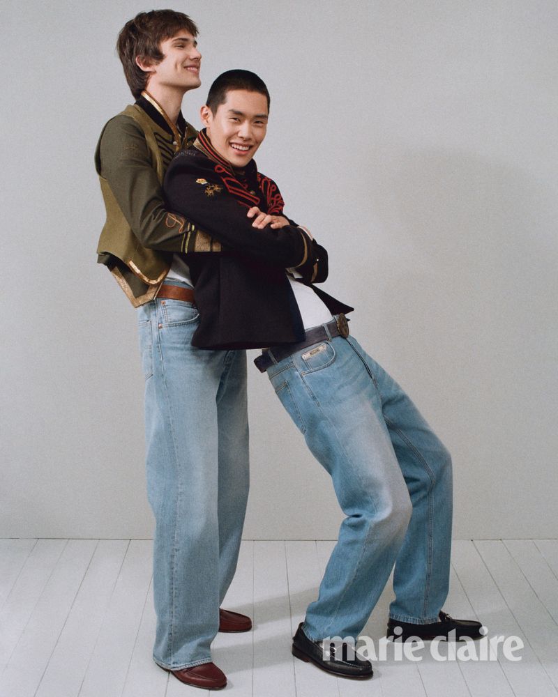Two male models smiling in military-style embroidered jackets and baggy light-wash denim jeans posing in a bright studio setting.