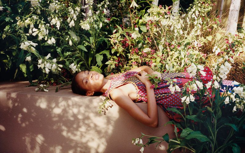 Model Chloe Paredes reclines on a sunlit ledge surrounded by blooming white foxgloves in a lush garden setting.