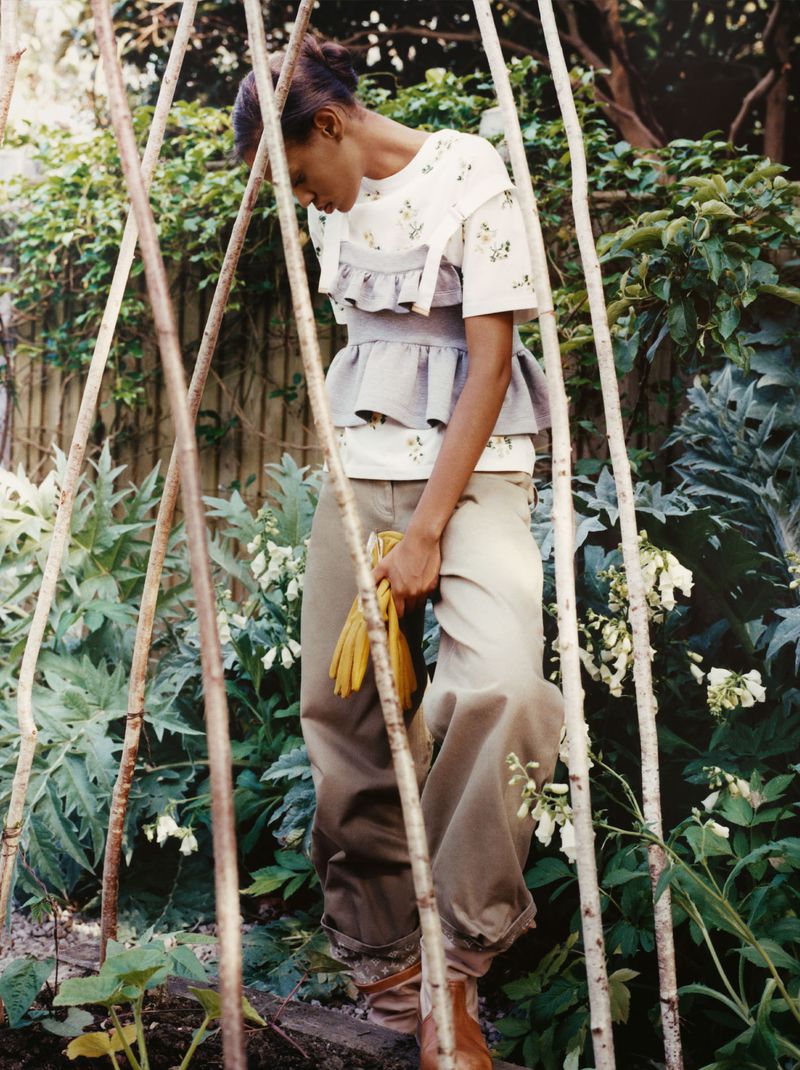 Model Chloe Paredes posing in layered ruffled top and wide beige trousers among bamboo garden stakes in a lush garden.