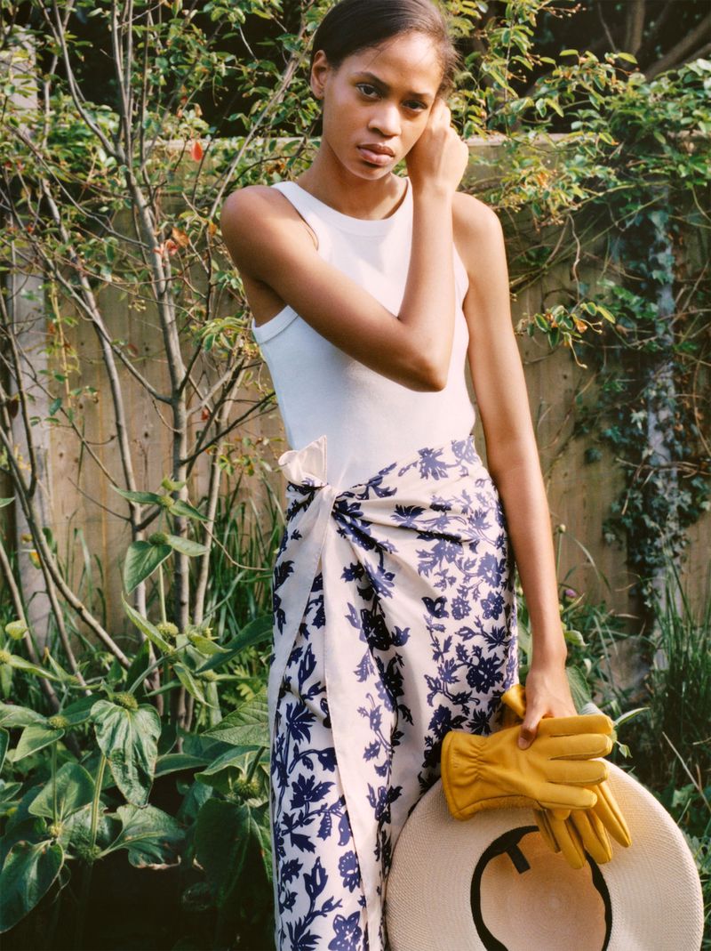 Model Chloe Paredes in a white tank top and floral wrap skirt holding yellow gloves and a straw hat in a garden.