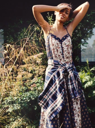 Model Chloe Paredes poses outdoors wearing a floral slip dress with a plaid shirt tied around her waist in a sunny lush garden.